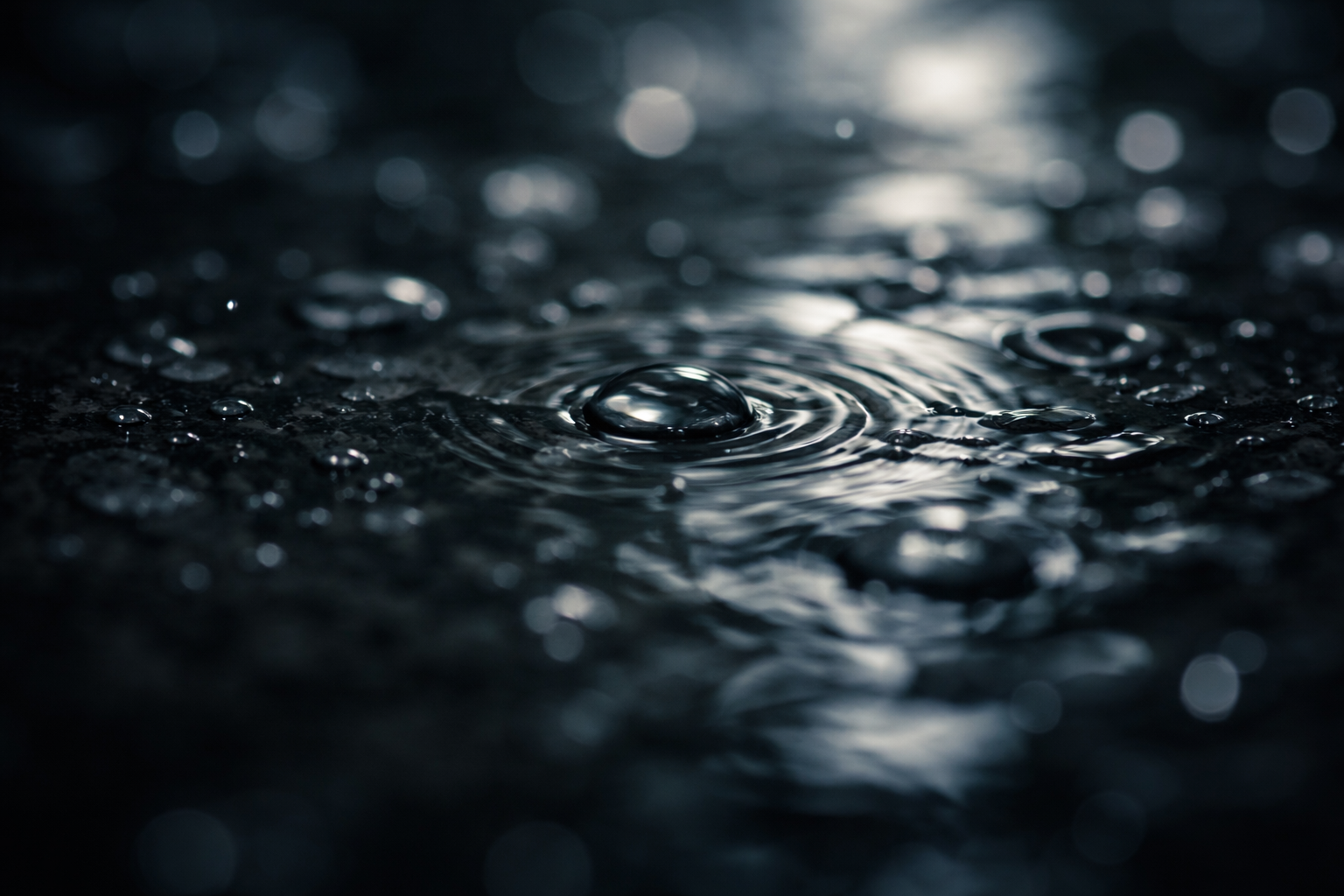 Macro close-up of water droplets and ripples on a dark reflective surface, with soft light creating abstract, blurred reflections.