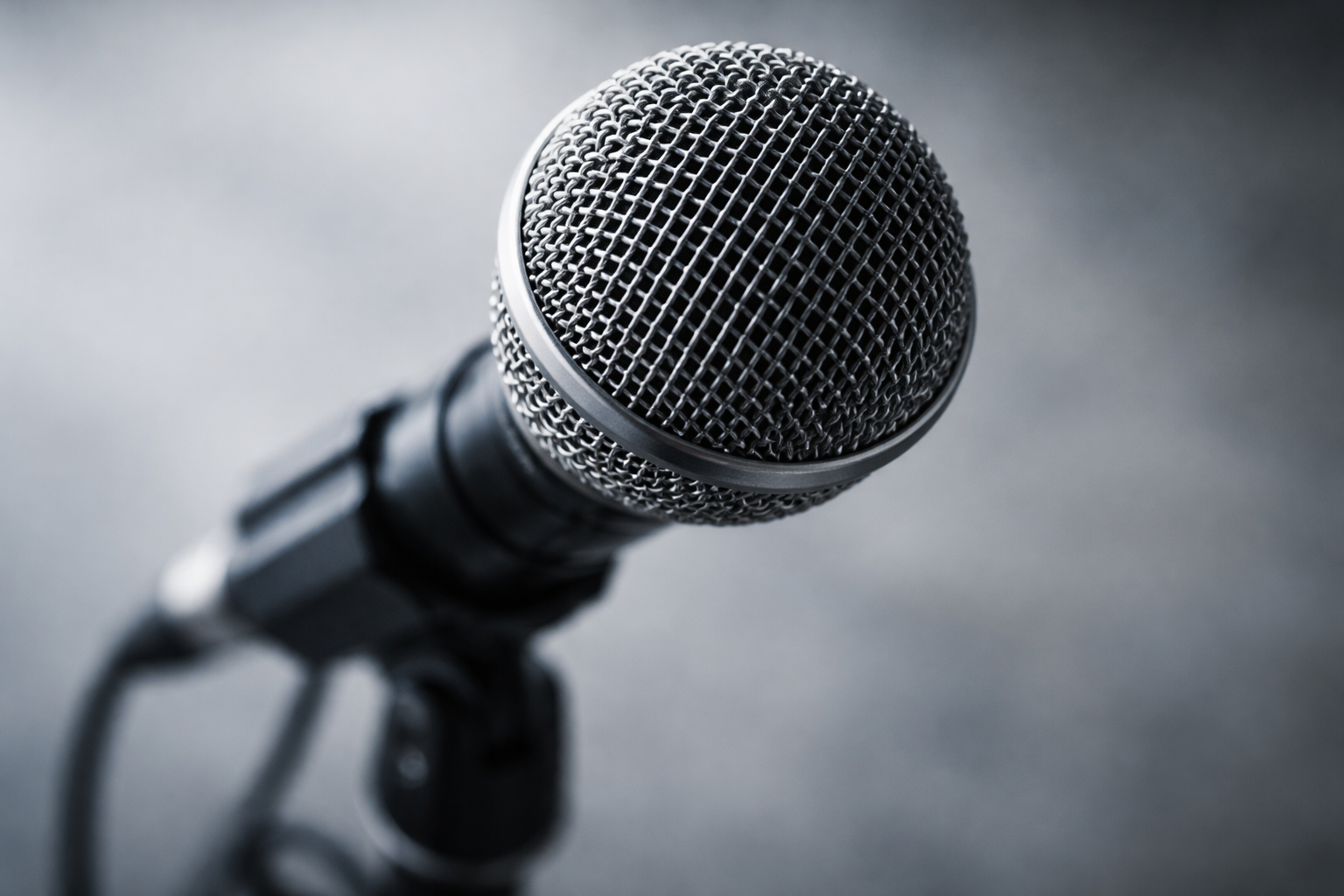 A close-up photograph of a microphone grille and stand, lit with soft overhead light, with an unfocused neutral background below.